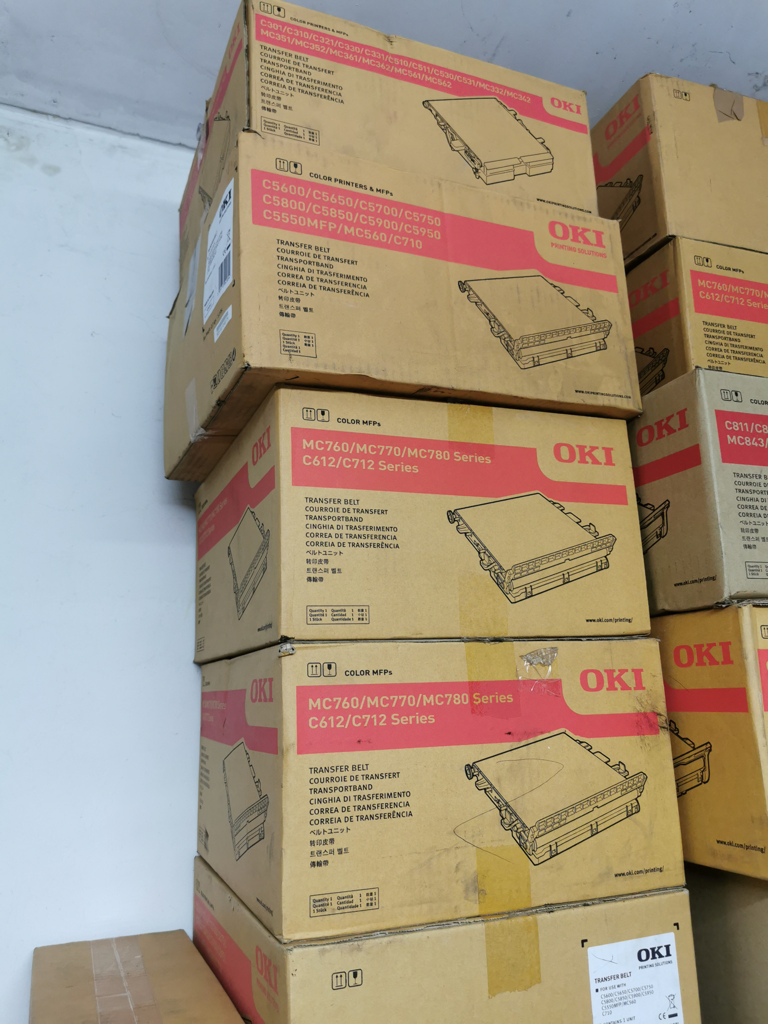 Stos kartonowych pudełek OKI z transfer beltami do kolorowych drukarek i urządzeń wielofunkcyjnych, modele C5600, MC760, C612. Widoczne napisy w kilku językach.