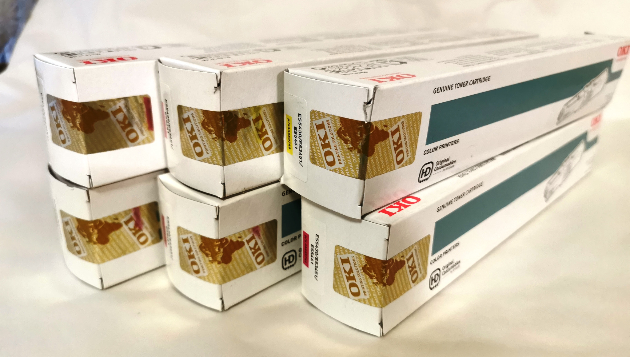 Stos pudełek z tonerami OKI do drukarek kolorowych, ułożonych w piramidę na jasnym tle. Widoczne etykiety z logo i oznaczeniami modelu.