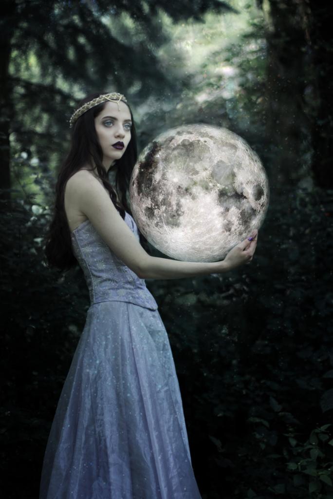 Kobieta w koronie i fioletowej sukni trzymająca w dłoniach świecącą kulę księżyca na tle ciemnego lasu.
