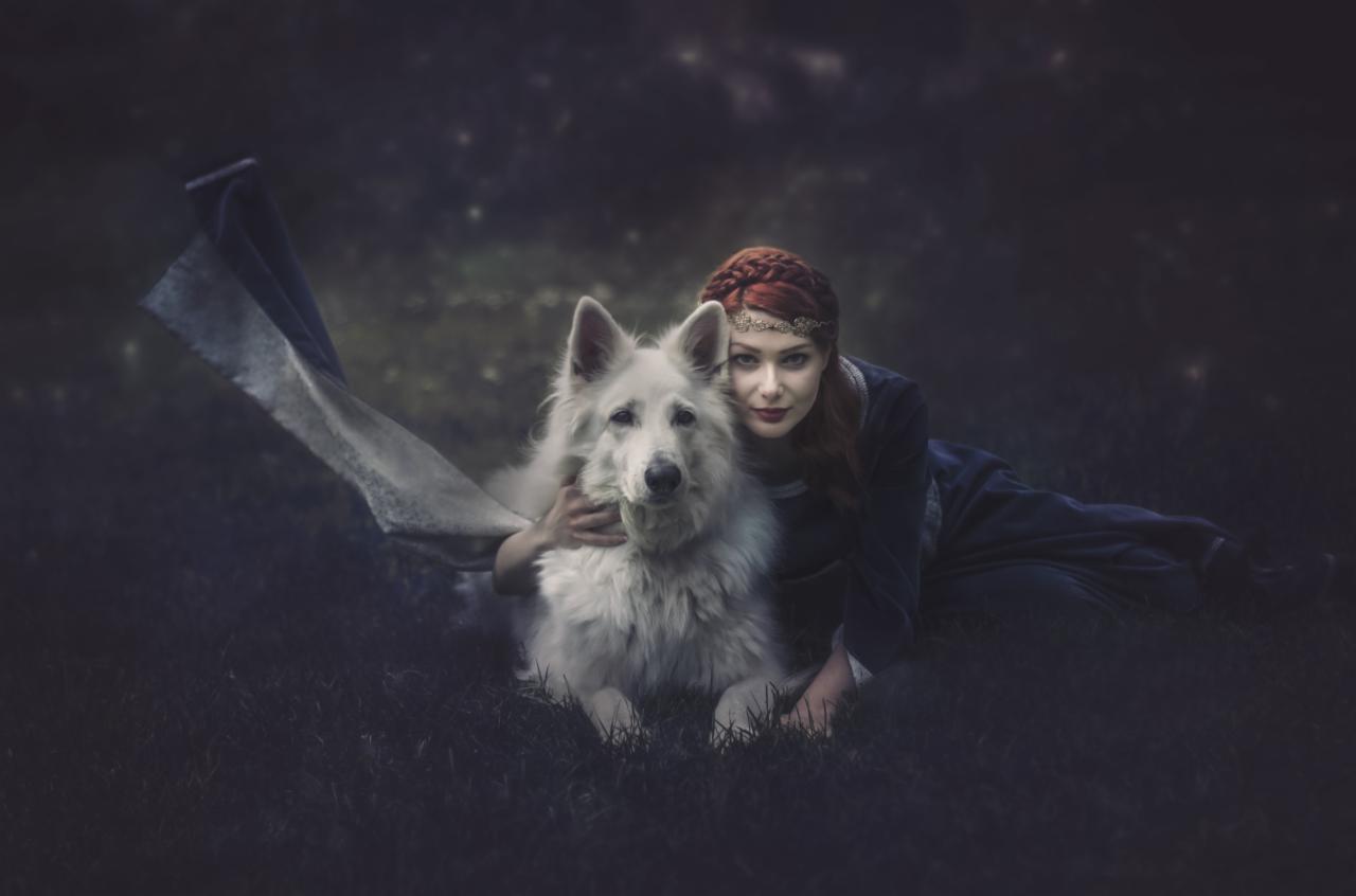 Kobieta z rudymi warkoczami w średniowiecznej sukni obejmuje białego psa na tle ciemnego, rozgwieżdżonego nieba.