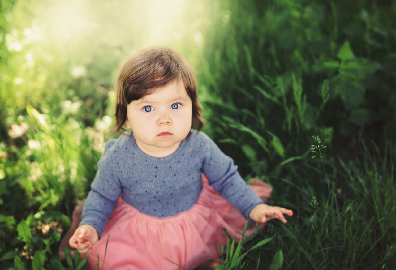 Urocza dziewczynka z niebieskimi oczami w szarej bluzce i różowej tiulowej spódniczce siedzi w wysokiej trawie w słoneczny dzień.