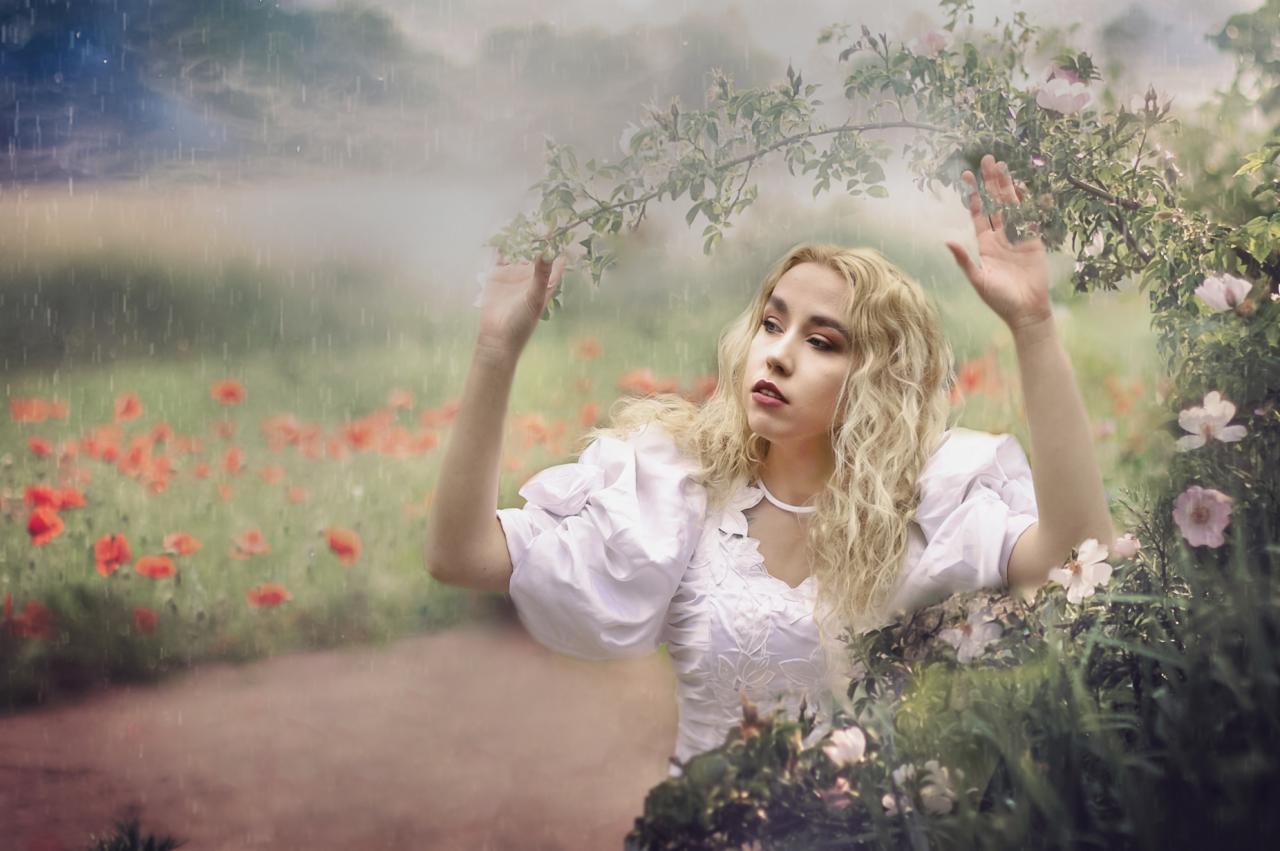 Portret kobiety w białej sukience, z blond włosami, stojącej w ogrodzie podczas deszczu, z ręką uniesioną ku gałęzi z kwiatami.