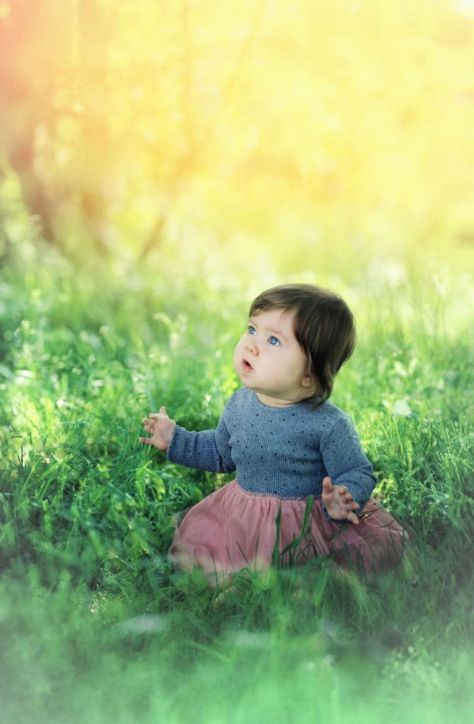 Niemowlę siedzi w wysokiej trawie, ubrane w szary sweter i różową tiulową spódniczkę, patrzy w górę na rozproszone światło słoneczne.