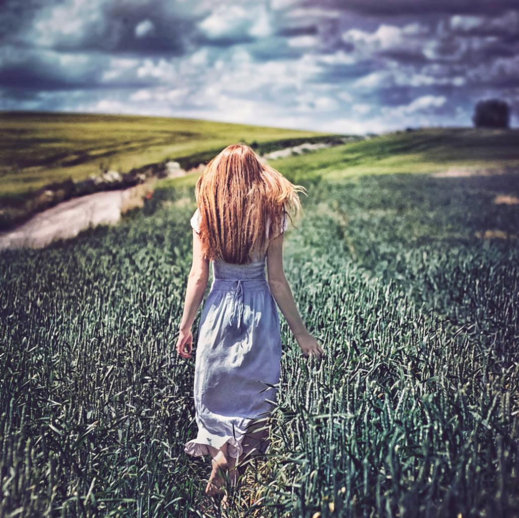 Kobieta w zwiewnej sukience idzie boso przez pole zboża, plecami do widza, z długimi rudymi włosami opadającymi na ramiona, w tle zielone wzgórza i pochmurne niebo.