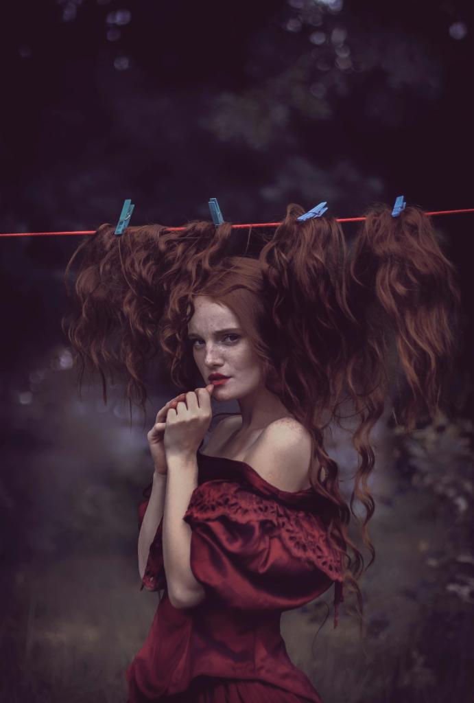 Portret rudej kobiety w burgundowej sukni, z trzema doczepianymi pasmami rudych włosów wiszącymi na sznurku do bielizny przypiętymi klamerkami, na tle ciemnego lasu.