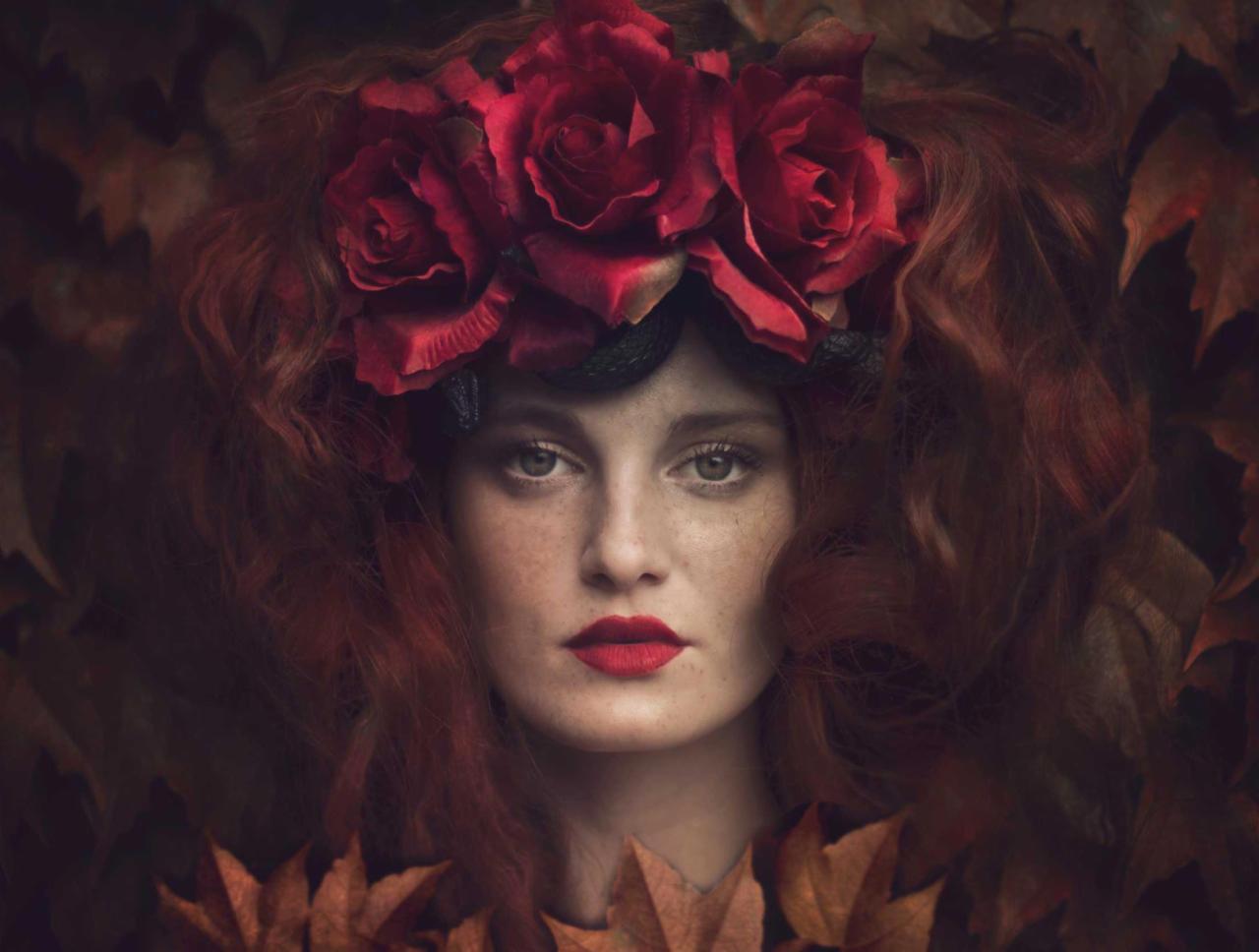 Portret kobiety o rudych włosach z wiankiem z czerwonych róż na głowie, na tle jesiennych liści w ciemnej tonacji.