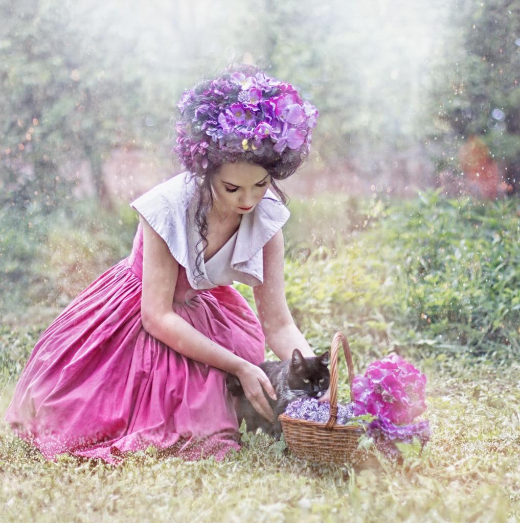 Kobieta w różowej sukience i kwiatowym nakryciu głowy kuca na trawie obok koszyka z kwiatami i czarnym kotem, bajkowa sceneria z rozmytym tłem.