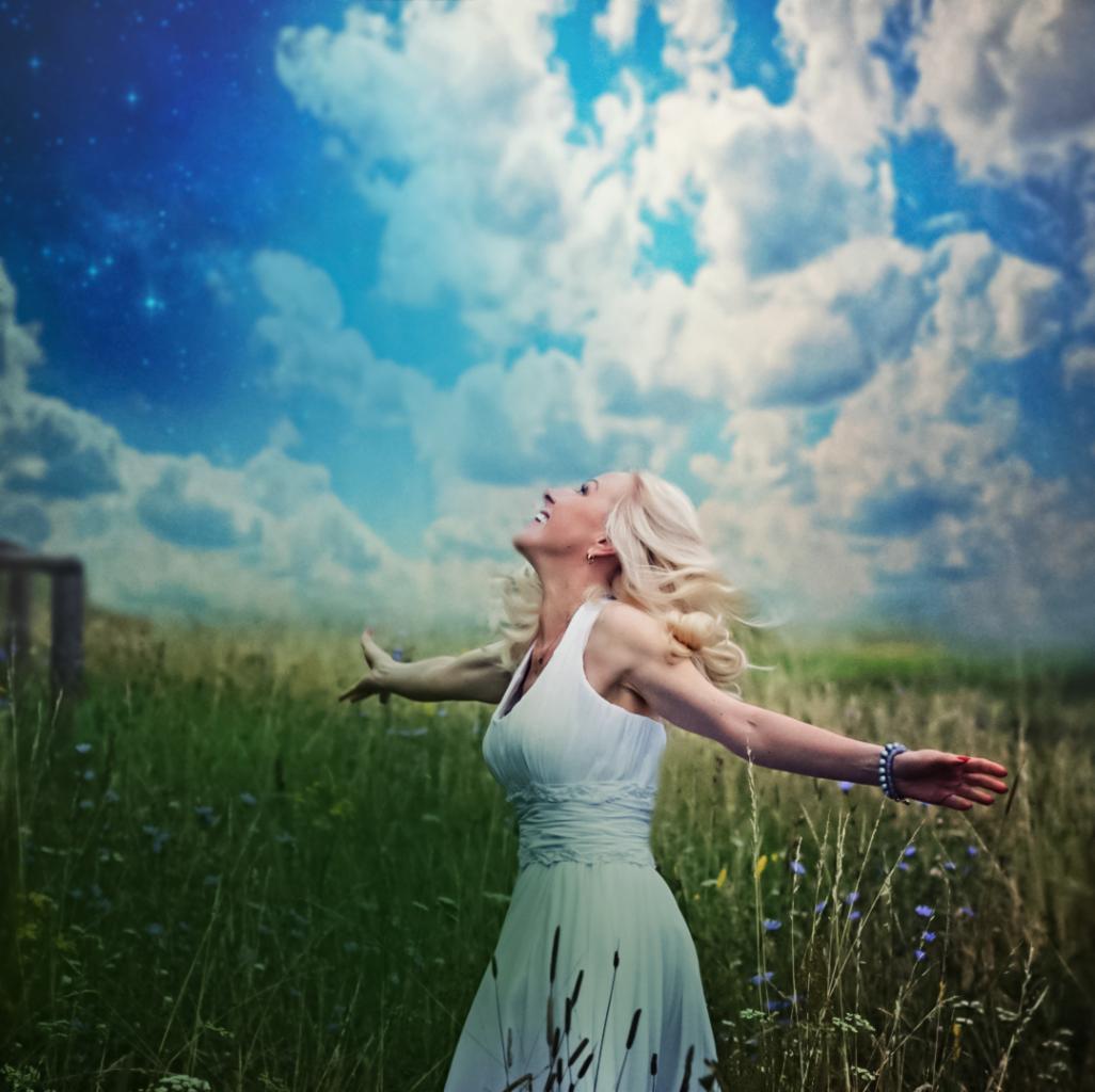 Kobieta w białej sukience, stojąca na łące z rozłożonymi ramionami, spogląda w górę na błękitne niebo z chmurami i gwiazdami, włosy rozwiane na wietrze.
