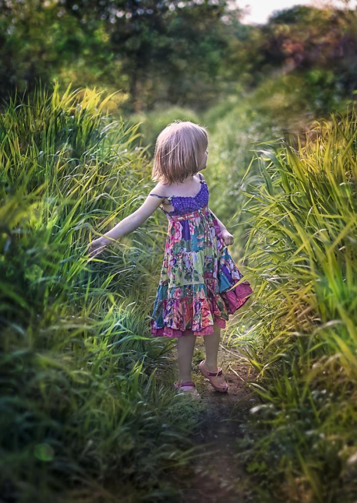 Dziewczynka w kolorowej sukience spaceruje ścieżką przez wysoką trawę, delikatnie dotykając źdźbeł dłonią, w tle rozmyty krajobraz.