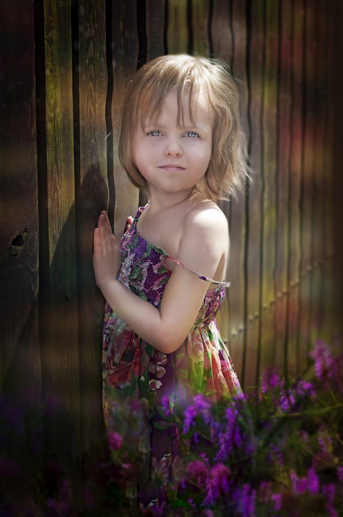 Dziewczynka w kwiecistej sukience opiera się o drewniany płot, otoczona fioletowymi kwiatami, z delikatnym uśmiechem na twarzy.