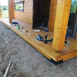 Drewniany taras w trakcie budowy przy domku letniskowym, widoczne narzędzia: wiertarka, piła tarczowa, poziomica. Świeżo położona drewniana podłoga tarasu. Piasek wokół tarasu.