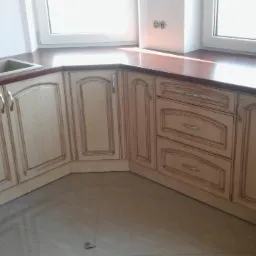 Jasna, narożna zabudowa kuchenna z frezowanymi frontami i drewnianym blatem, widok z dołu, jasne płytki na podłodze.