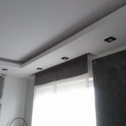 Nowoczesny salon z podwieszanym sufitem, wpuszczonymi lampami, szarą zasłoną i ścianą w odcieniach szarości.