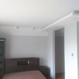 Jasne wnętrze sypialni po odświeżeniu z pomalowanymi ścianami na szaro, z zabudową nad łóżkiem i oświetleniem punktowym na suficie.