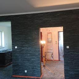 Usługi remontowo-budowlane - Montaż dekoracyjnej ściany z czarnego kamienia łupkowego w nowym wnętrzu, widoczne przygotowania do dalszych prac wykończeniowych, w tle drabina i lampa budowlana.