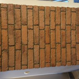 Usługi remontowo-budowlane - Ściana wyłożona czerwoną cegłą klinkierową, ułożoną pionowo, z widocznymi fugami, fragment okna i sufitu w tle, dolna krawędź ściany wykończona listwą.