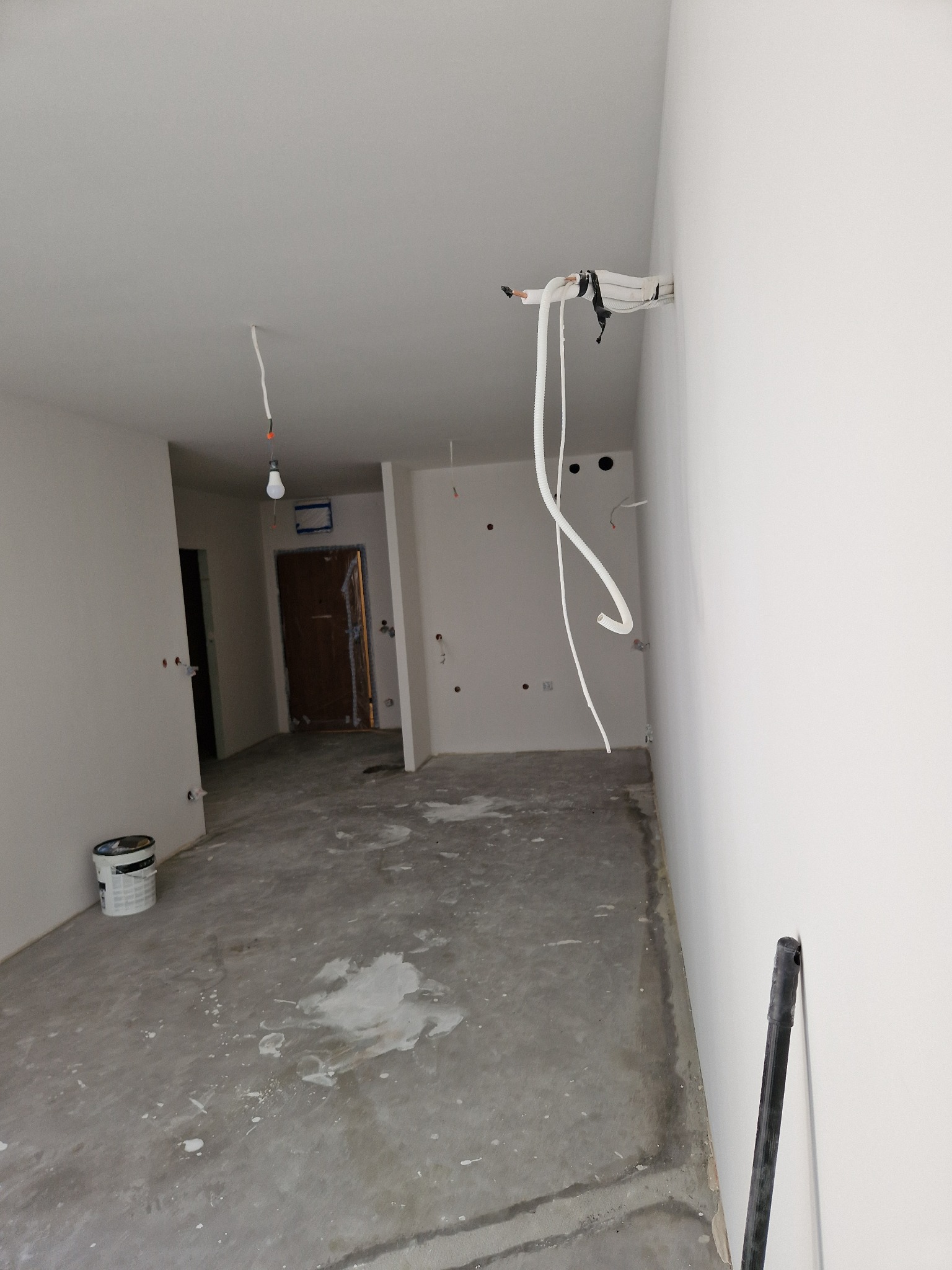 Wnętrze w trakcie remontu: surowe ściany, wystające kable, ślady farby na podłodze i wiadro z farbą. Widoczne przygotowania do prac wykończeniowych.