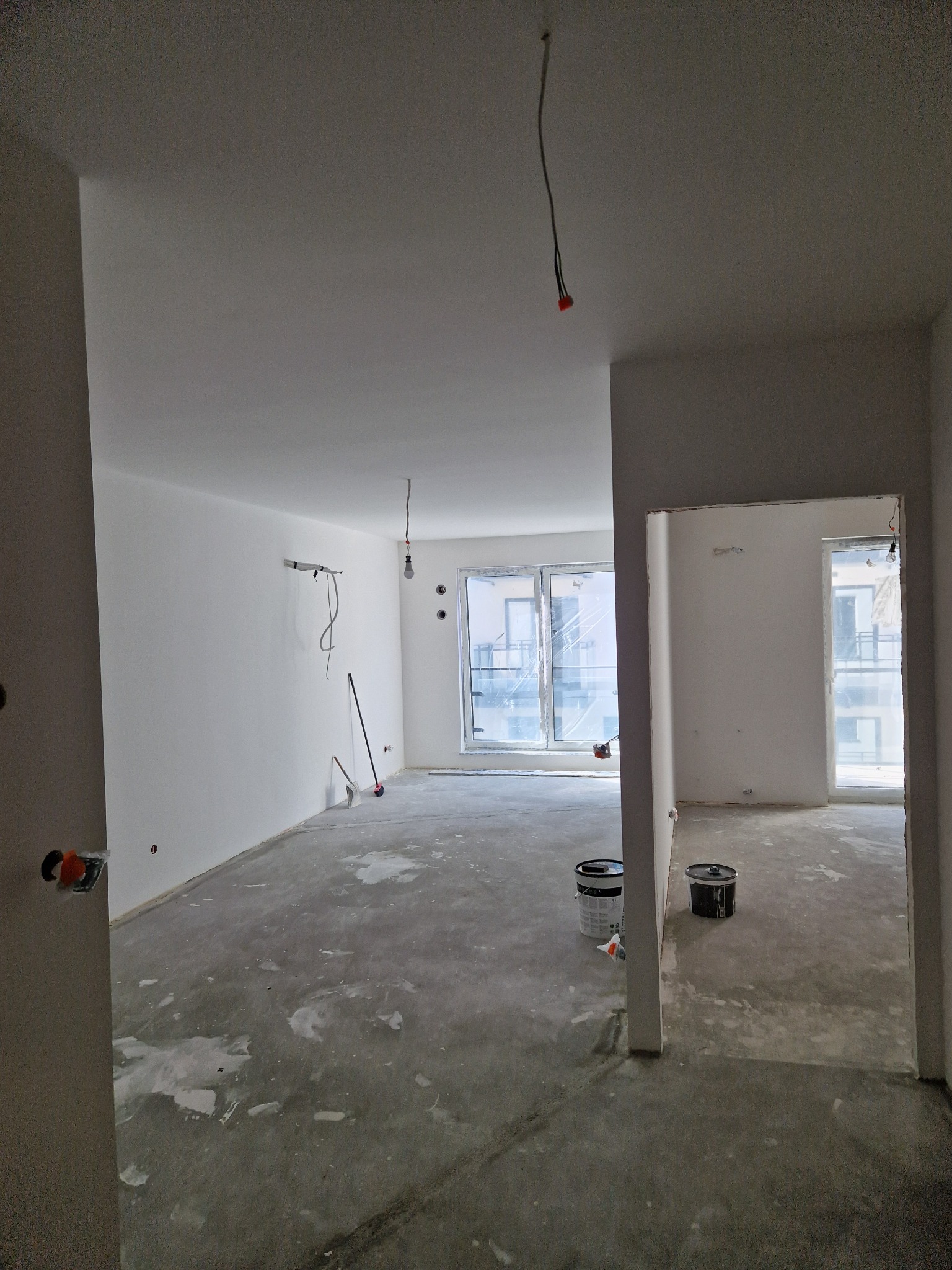 Wnętrze po szpachlowaniu ścian na biało, widoczne puszki z farbą i wystające przewody elektryczne. Duże okna z widokiem na zewnątrz.