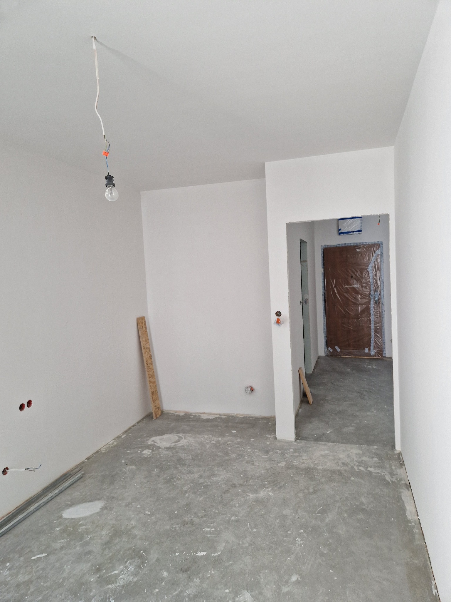 Wnętrze w trakcie prac wykończeniowych: surowe ściany pomalowane na biało, widoczne przewody elektryczne, zabezpieczone drzwi wejściowe. Podłoga surowa, przygotowana do dalszych prac.