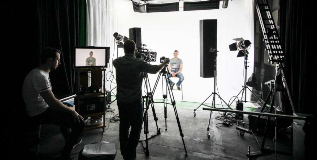 Studio filmowe z kamerzystą filmującym mężczyznę siedzącego na krześle na białym tle, podgląd na monitorze, oświetlenie studyjne.