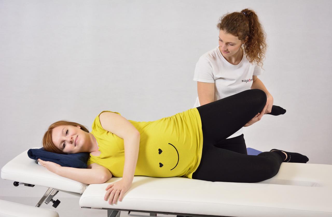 Kobieta leży na kozetce, ubrana w żółtą koszulkę z nadrukiem uśmiechniętej buźki, podczas gdy fizjoterapeutka w białej koszulce delikatnie unosi jej nogę, wykonując ćwiczenie rehabilitacyjne.