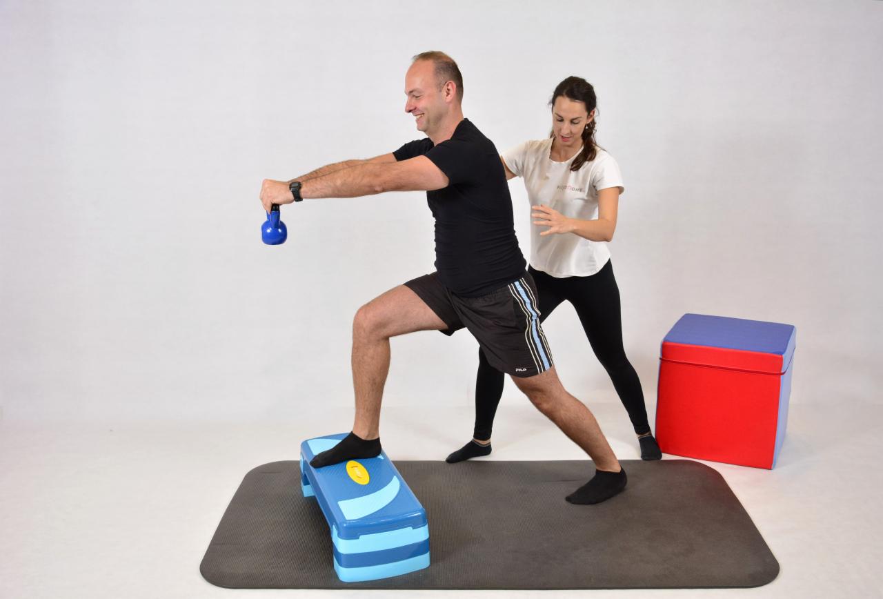 Mężczyzna wykonuje wykrok z obciążeniem (niebieski kettlebell) na stepie, pod nadzorem kobiety w białej koszulce.