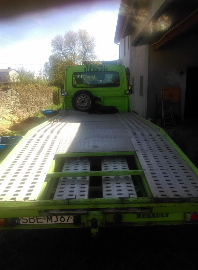 Zielona laweta Renault z numerem telefonu na kabinie, zaparkowana obok budynku z drabiną.