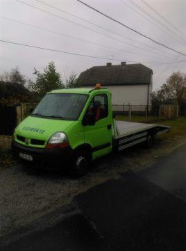 Jaskrawozielony samochód typu laweta Renault Master na żwirowym podłożu przed białym budynkiem z ciemnym dachem w pochmurny dzień. Pomarańczowe światło ostrzegawcze na dachu kabiny.