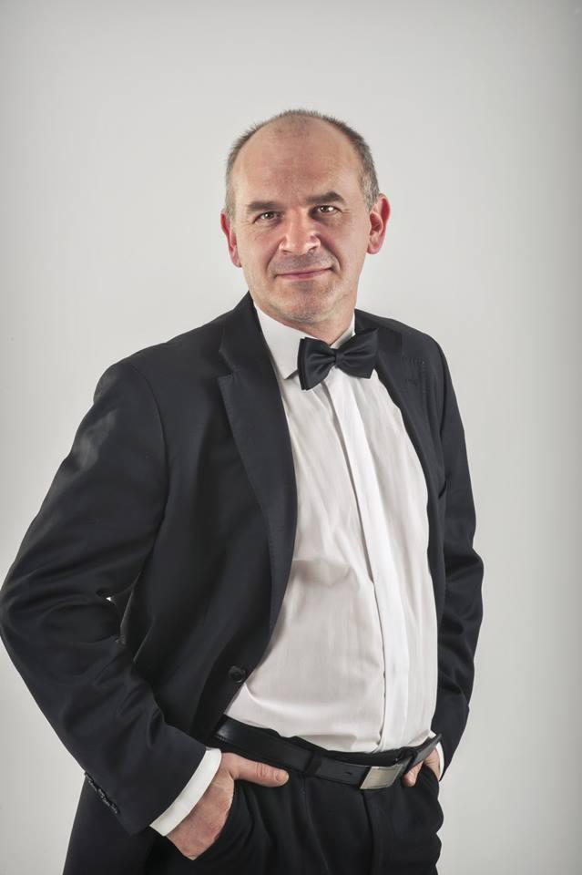 Portret mężczyzny w czarnym smokingu i muszce na jasnym tle, z ręką w kieszeni.