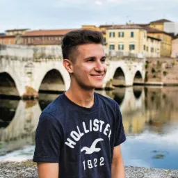 Uśmiechnięty młody mężczyzna w koszulce Hollister na tle kamiennego mostu z odbiciem w wodzie.