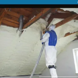 Pracownik w kombinezonie ochronnym aplikuje piankę izolacyjną na poddaszu, widoczne drewniane elementy konstrukcji dachu i przewód wentylacyjny.