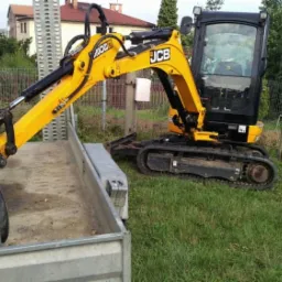 Żółta minikoparka JCB na trawie, łyżka załadowana ziemią na przyczepie, w tle ogrodzenie i domy.