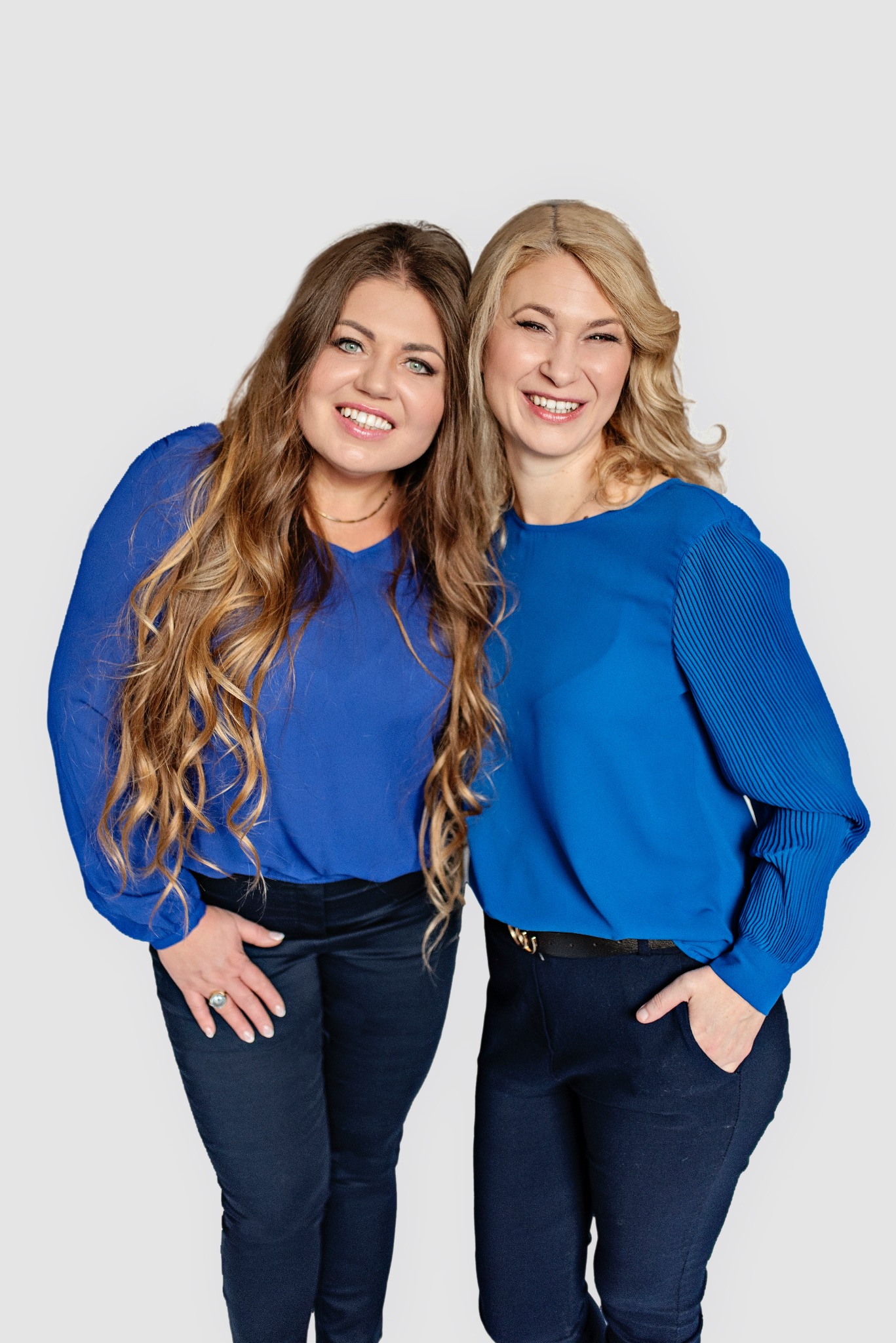 Dwie uśmiechnięte kobiety w błękitnych bluzkach, prezentujące się na jasnym tle, jedna z ręką w kieszeni, druga z dużym pierścieniem na palcu.