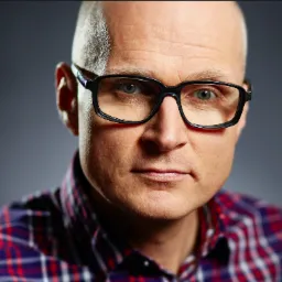 Portret łysiejącego mężczyzny w okularach o czarnych oprawkach i koszuli w kratę na szarym tle.