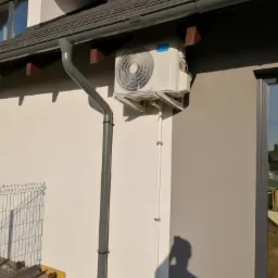 Zewnętrzna jednostka klimatyzacji Midea zamontowana na ścianie budynku, widoczne rury odprowadzające skropliny i przewody elektryczne, cień osoby robiącej zdjęcie na ścianie.