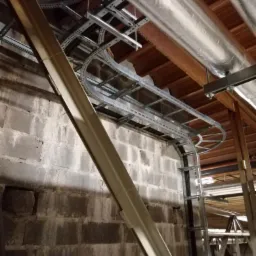 Metalowe korytka kablowe zamontowane pod sufitem w pomieszczeniu z surową, ceglaną ścianą i widocznymi rurami wentylacyjnymi.