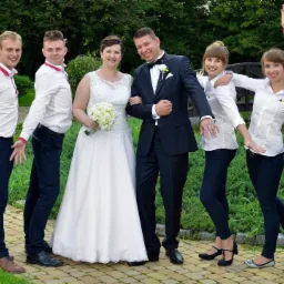 Para młoda pozuje do zdjęcia z grupą osób w identycznych strojach z logo firmy na koszulach, na tle fontanny i zieleni.