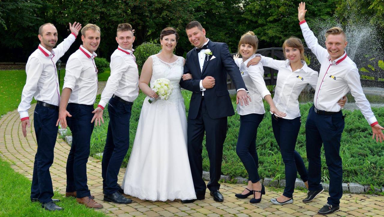 Para młoda pozuje do zdjęcia z grupą osób w identycznych strojach z logo firmy na koszulach, na tle fontanny i zieleni.