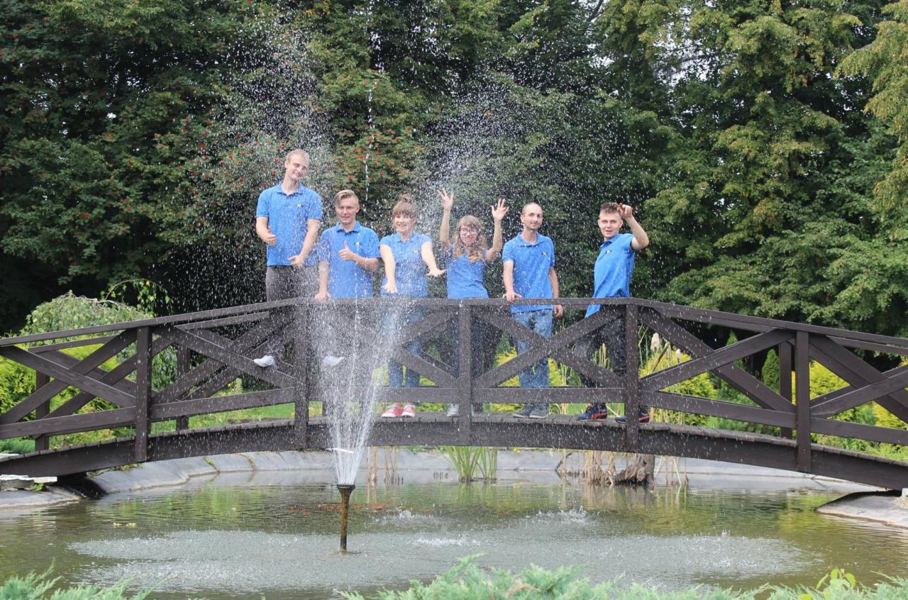 Sześcioosobowa grupa w niebieskich koszulkach pozuje na drewnianym mostku nad stawem z fontanną, otoczonym zielenią