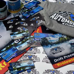 Zestaw gadżetów reklamowych Automobilklubu w Przemyślu: kubki, wizytówki, ulotki i szara koszulka z nadrukowanym logo i nazwą klubu oraz hasłem 'Drift Przemyśl 2021 Królowie Ulicy'.