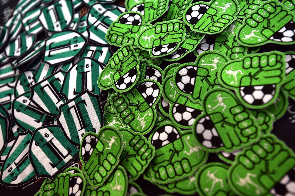 Stos zielonych i białych naszywek: zielone pięści z piłką nożną i piłkarzem oraz tarcze z literą 'T' i rokiem 1945, widoczne detale haftu i tekstury materiałów.