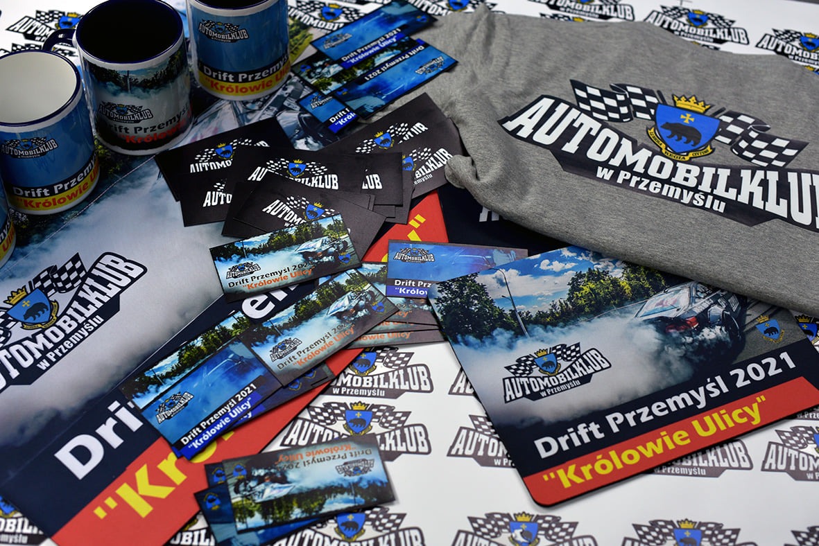 Zestaw gadżetów reklamowych Automobilklubu w Przemyślu: kubki, wizytówki, ulotki i szara koszulka z nadrukowanym logo i nazwą klubu oraz hasłem 'Drift Przemyśl 2021 Królowie Ulicy'.