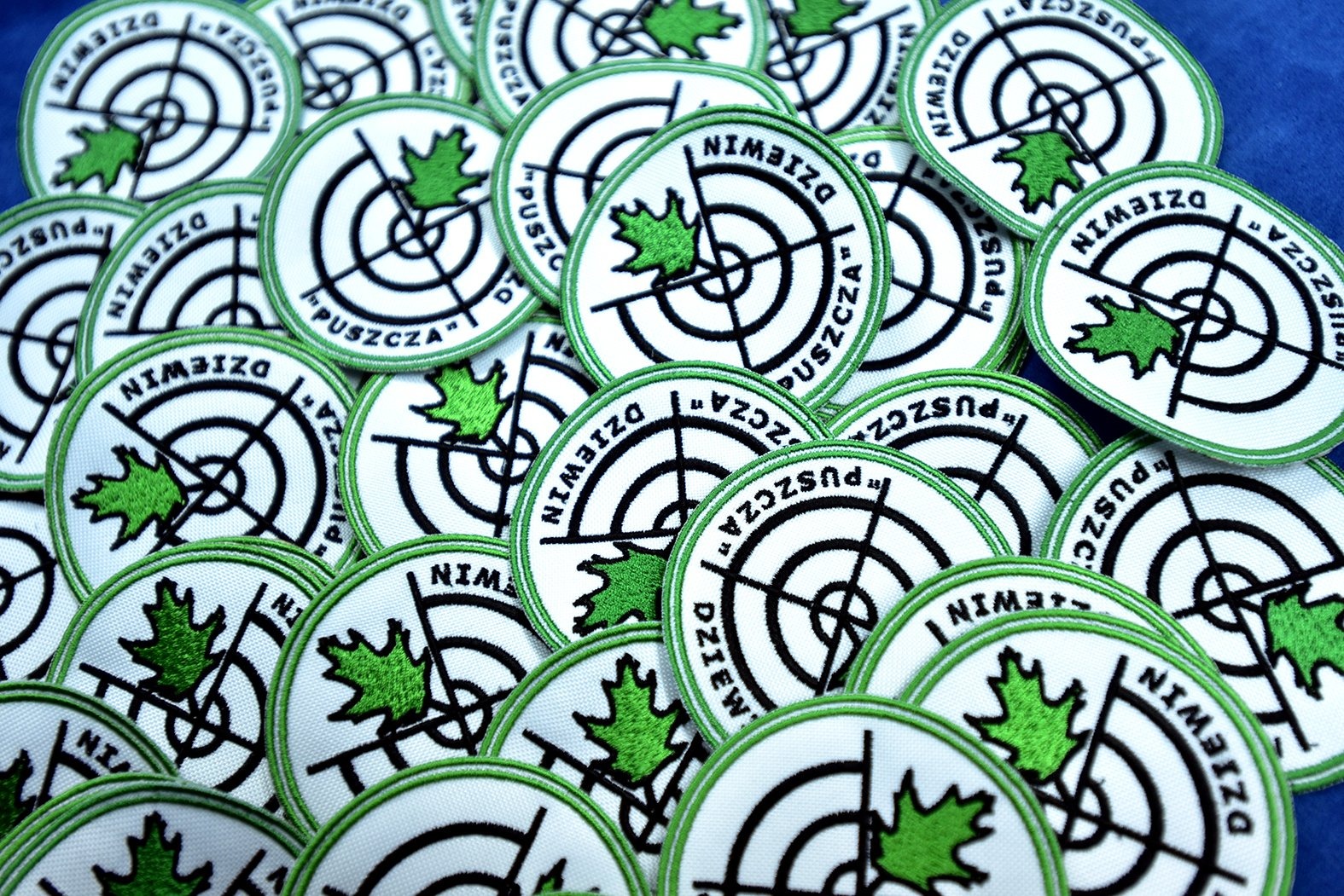 Stos okrągłych naszywek z białym tłem, zieloną obwódką i motywem tarczy strzelniczej z zielonym liściem dębu pośrodku oraz napisem 'DZIEWIN PUSZCZA'