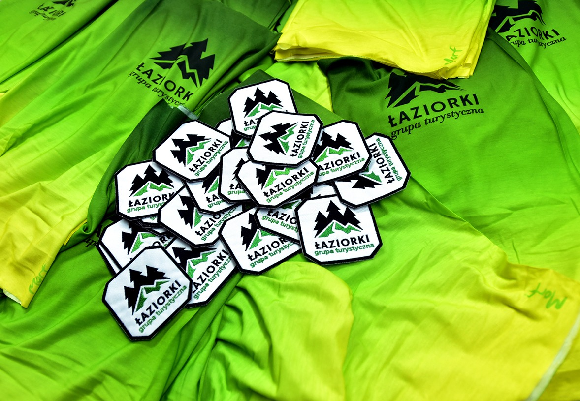 Stos zielono-żółtych koszulek z nadrukowanym logo firmy turystycznej oraz naszywki z tym samym logo leżące na wierzchu, całość sfotografowana z góry.