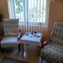 Pokój z dwoma fotelami obitymi materiałem w zygzaki, stolikiem nakrytym białym obrusem i oknem z żaluzjami pionowymi.