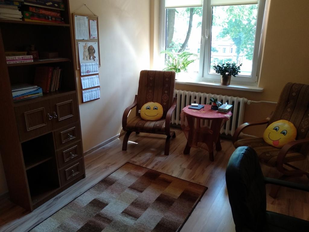 Gabinet z dwoma fotelami z poduszkami w kształcie emotikonów, mały stolik z notatnikiem i długopisem, regał z książkami i kalendarz na ścianie, dywan w geometryczne wzory.