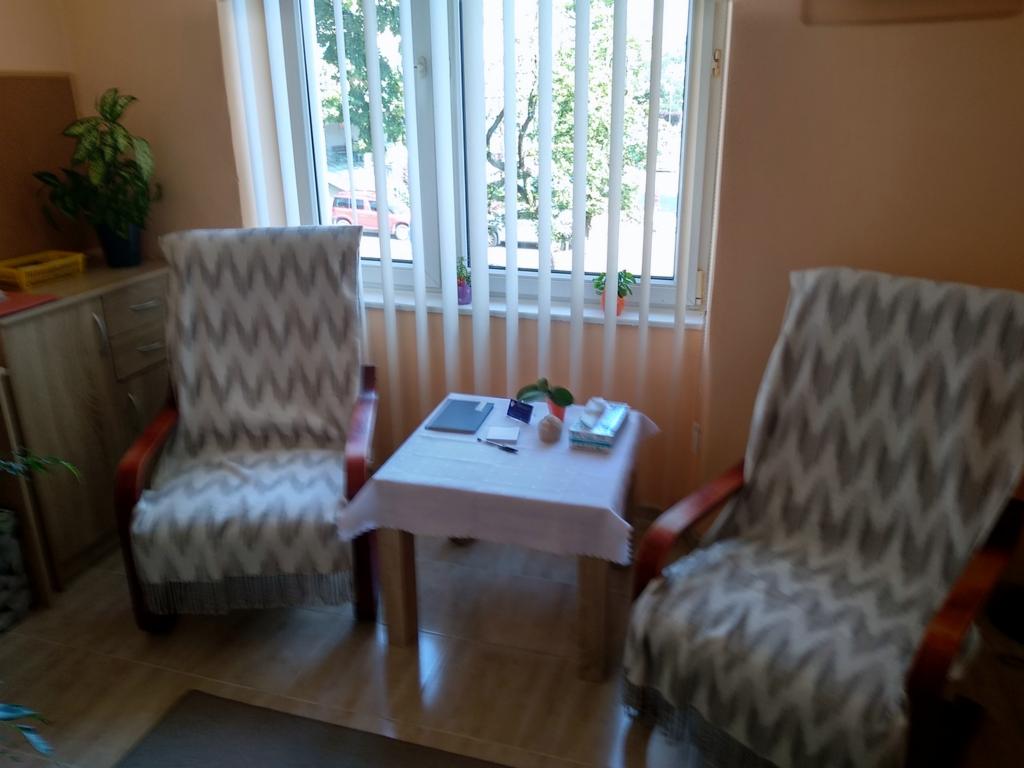 Pokój z dwoma fotelami obitymi materiałem w zygzaki, stolikiem nakrytym białym obrusem i oknem z żaluzjami pionowymi.