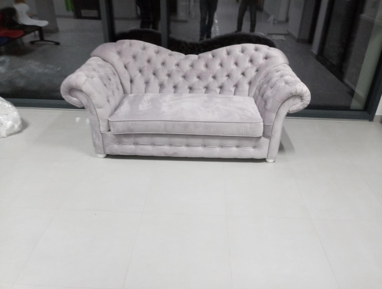 Jasnoszara sofa typu chesterfield z głębokimi pikowaniami, ustawiona na białej podłodze z płytek, widok z przodu.