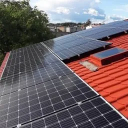 Panele słoneczne na czerwonym dachu z dachówkami, widoczne okno dachowe i fragment zieleni w tle.