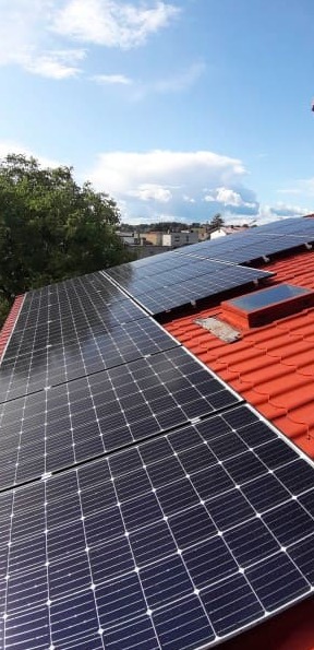 Panele słoneczne na czerwonym dachu z dachówkami, widoczne okno dachowe i fragment zieleni w tle.